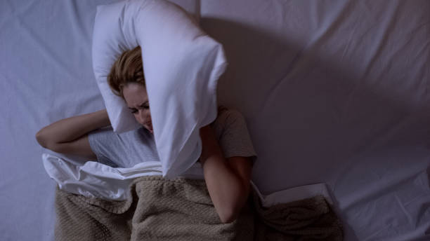 Angry girl closing ears with pillow, noisy neighbors disturbing sleep at night
