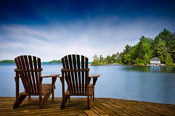 A couple of wooden Muskoka chairs sitting on the dock with a lake and cottages across in the background.
